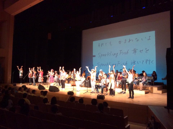 男女10名が舞台の上でスタンドマイクの前に立ち観客席に大きく手を広げ手拍子している写真