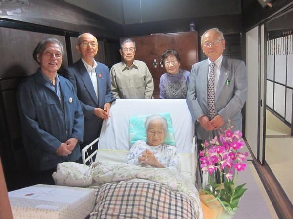 介護ベッドで起き上がっている前川たつさん(101歳)が長寿祝いの花と記念品を前に市長らと一緒に写っている写真