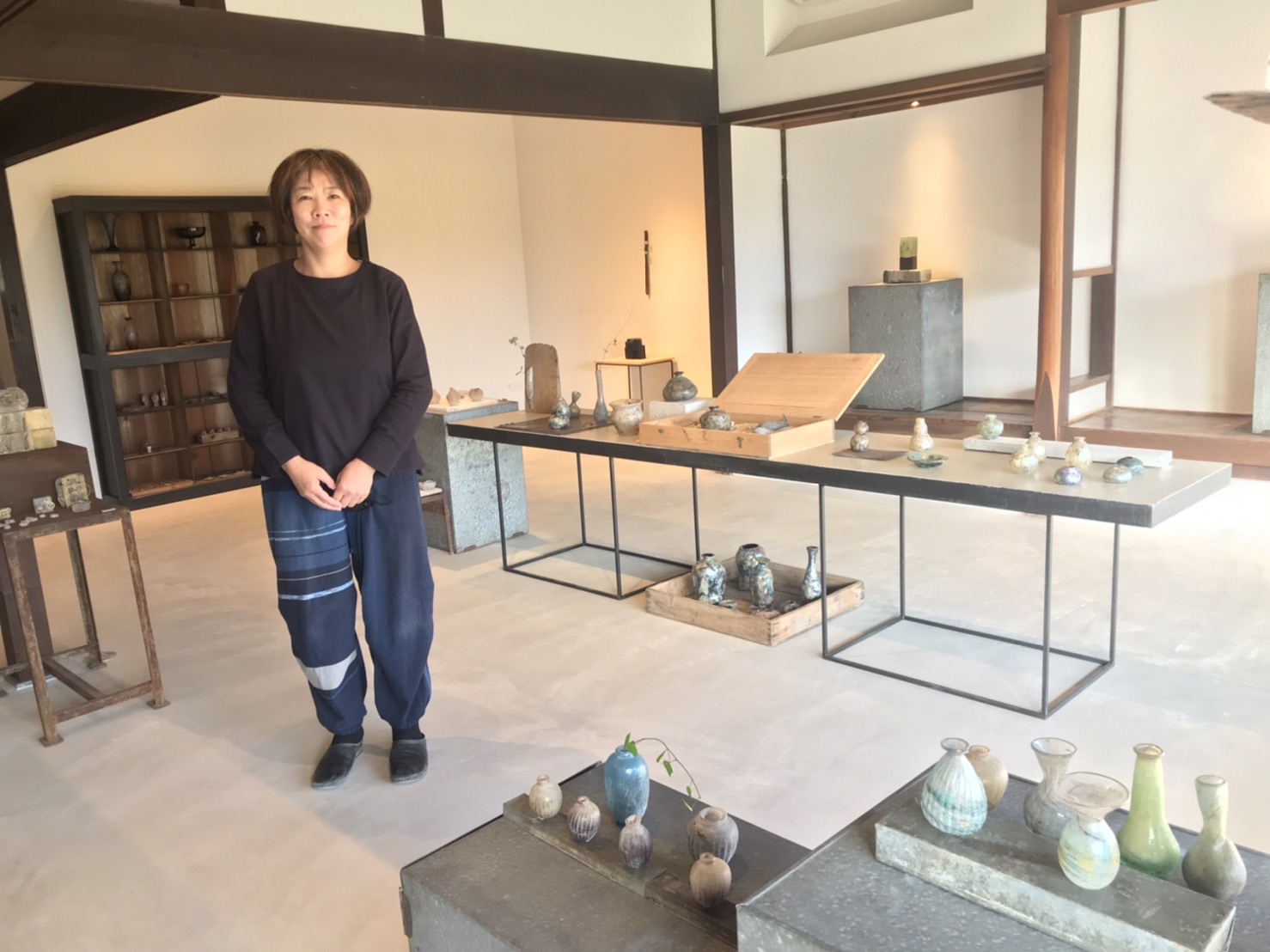 ガラスの作品を並べたギャラリー内に女性の職人が立っている