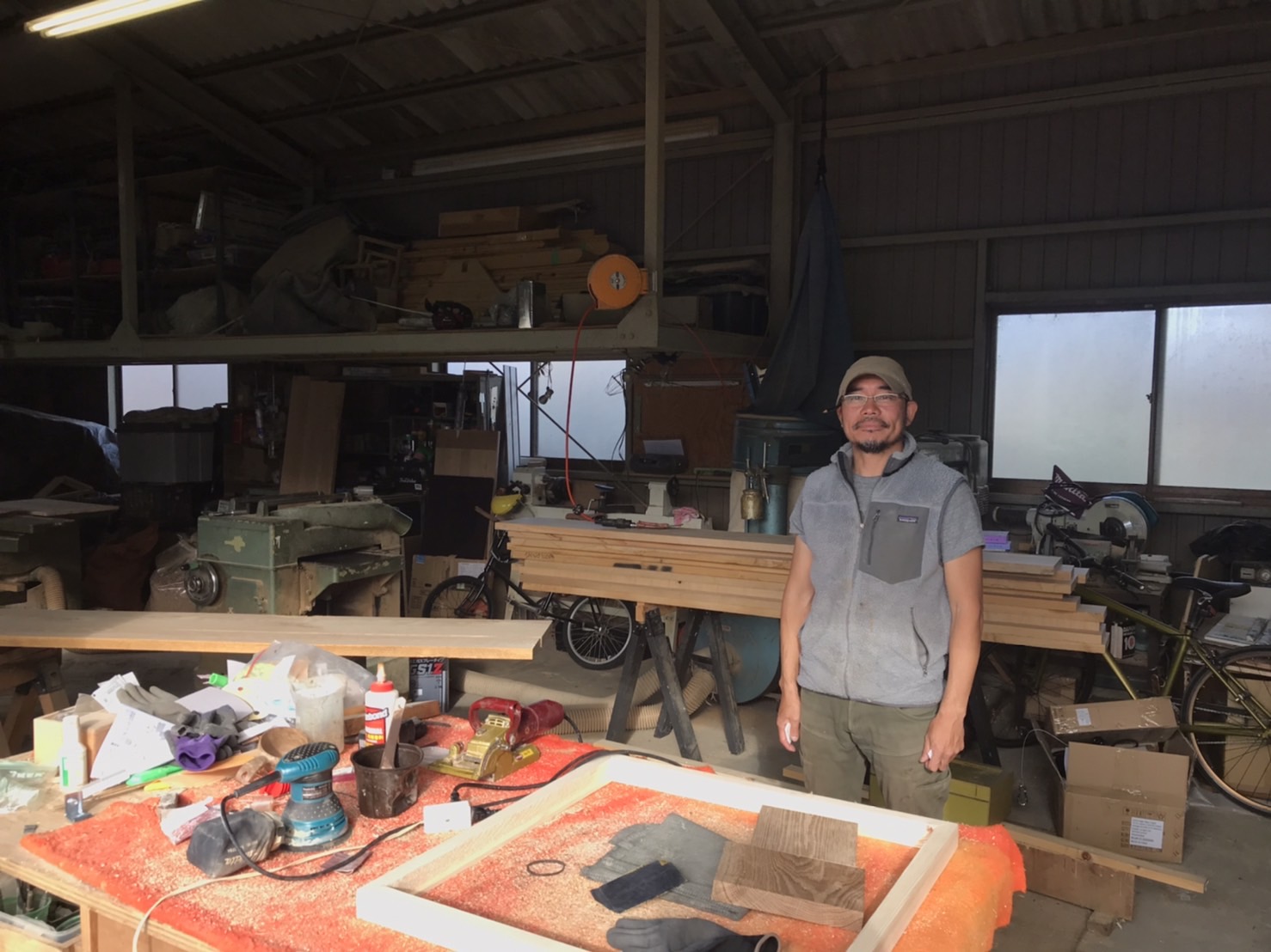 木材や工具が置いてある作業場で職人の男性が立っている