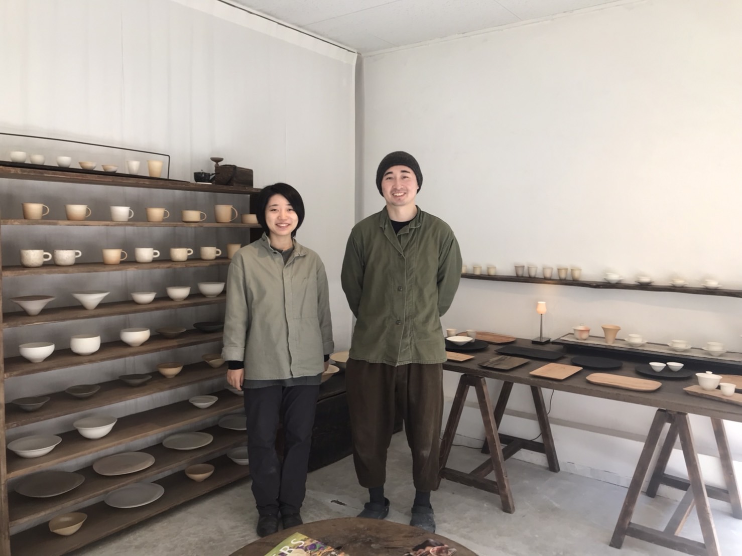 机と棚一面に作品のカップやお皿などが並んでいて男性と女性の職人が立っている