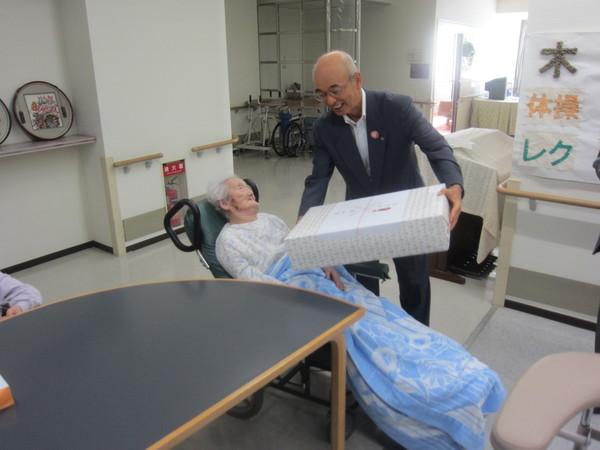車いすベッドに乗っている圓増艶子さん(104歳)が市長より長寿祝いの記念品を贈呈されている写真