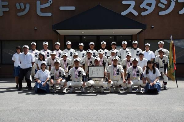 野球場の前で、優勝旗と賞状を見せて、首からメダルをかけた選手達と部員達も一緒に写っている記念撮影の写真