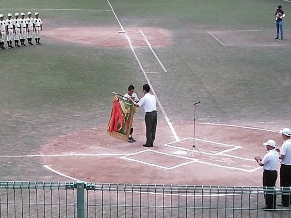 グランドのバッター席の前で、白いポロシャツを着た男性が、選手の一人に優勝旗を手渡ししている様子の写真