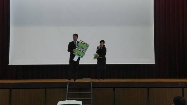 舞台で、背広を着た男性がパンフレットのような物を持ってマイクを持っている女性が話をしている様子の写真