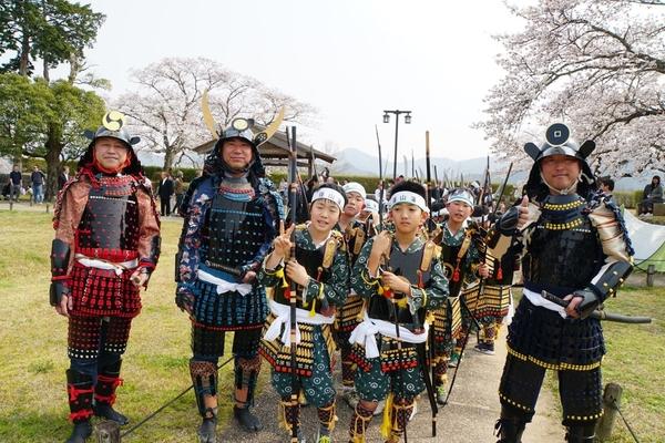 鎧兜を着た男性3名と鉢巻きに鎧姿の子供たちが槍を持って桜をバックに記念撮影している写真
