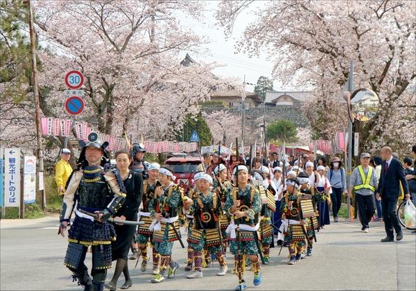 鎧兜を着た男性を先頭に鉢巻きに鎧姿の子供たちが槍を持って桜並木を練り歩いている写真