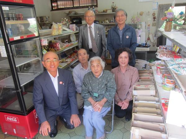 パイプ椅子に腰かけている長寿祝いの上田ふみこさん(101歳)が店内で市長らと記念撮影している写真