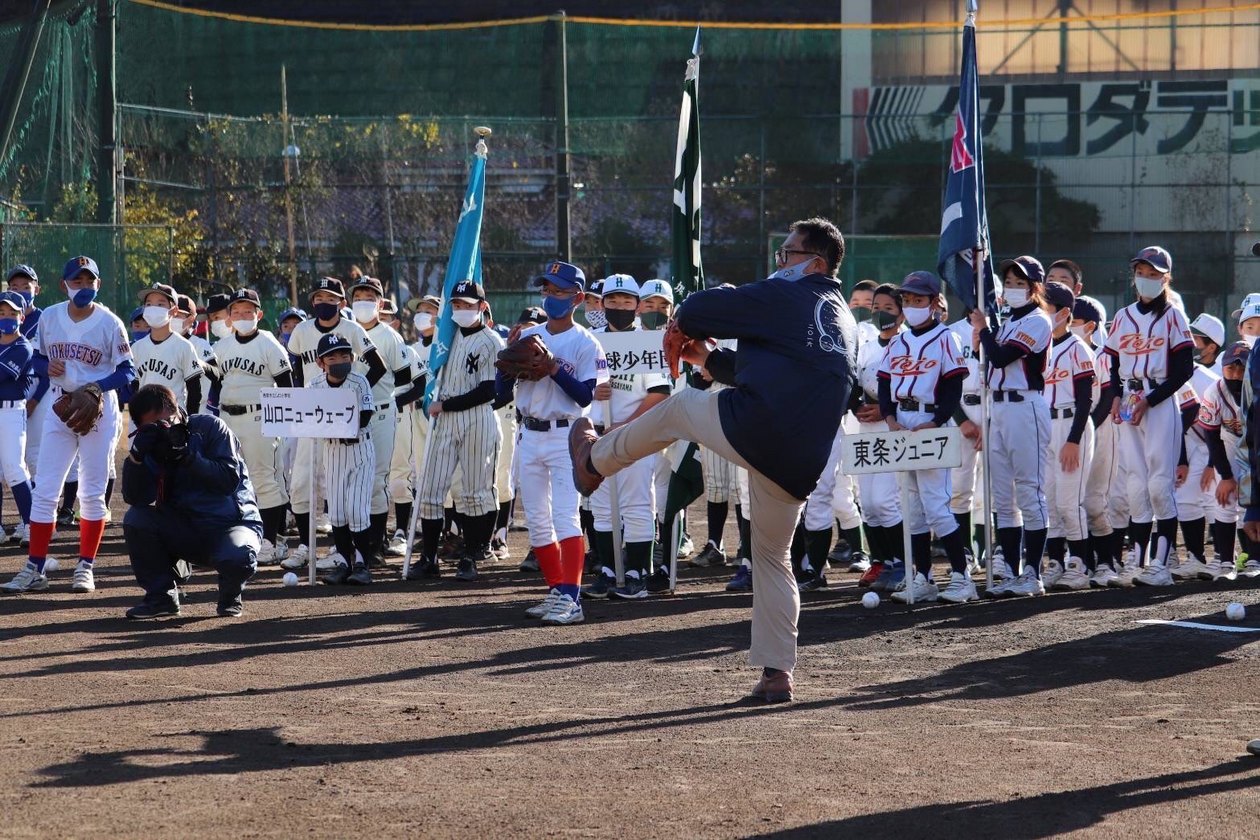 始球式で教育長がマウンドから投げようとされている。後ろで出場選手たちが見守っている。