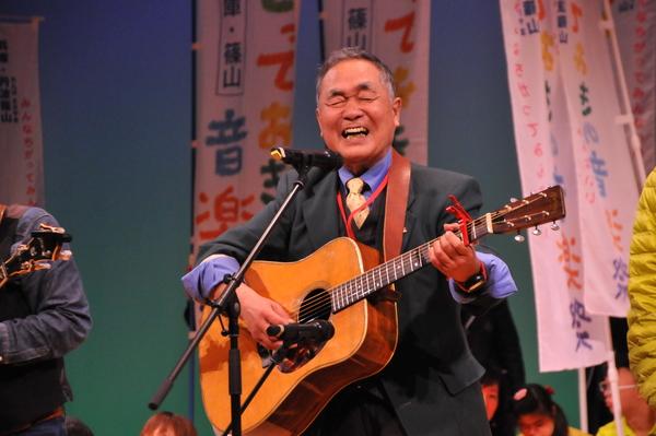 ホールの舞台で男性がギターを弾きながら熱唱している写真