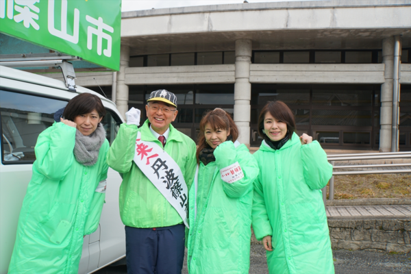 選挙カーの前で、黄緑色のジャンパーとタスキを付けて市長がてガッツポーズをして女性3人と映っている写真