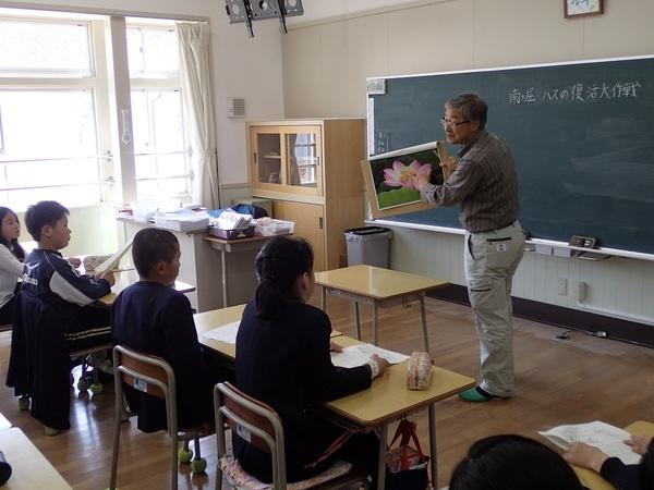 小学校の教室で、額に入ったハスの絵を持って生徒たちに説明をしている男性の写真