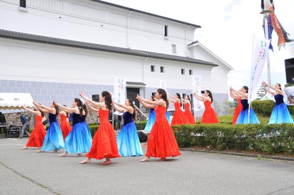 建物の外で鯉のぼりが風になびく中、赤色や青色の衣装を着た女性らがフラダンスを踊っている写真