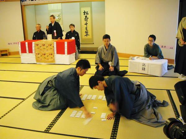 対戦中に札を取り合っている様子を見ている審判と記録している人が写っている写真