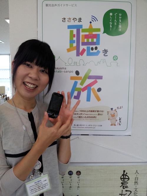 スマートフォンを手に持った女性の後ろの壁にささやま聴き旅のポスターが貼ってある写真