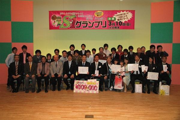 S－1グランプリの横断幕の前で、受賞した出場者が賞状を持って座っており、市長や関係者も一緒に写っている集合写真