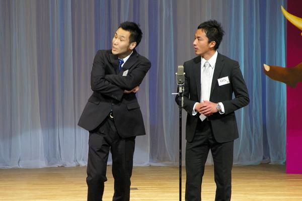 左側の男性が腕を組んでおり、右側の男性が両手を合わせている、「コンフィング」というグループが漫才をしている様子の写真