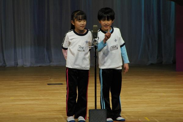 「ミックスツインズ」というグループ名の体操服を着ている小学生の男の子と女の子がスタンドマイクの前で漫才をしている写真