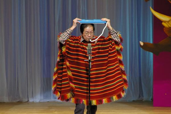 鍵盤ハーモニカのホースを口に加えて、鍵盤ハーモニカを両手で頭の上にのせ、シマシマ模様の羽織物を着ている「マエストロ足立」さんの漫才の様子の写真