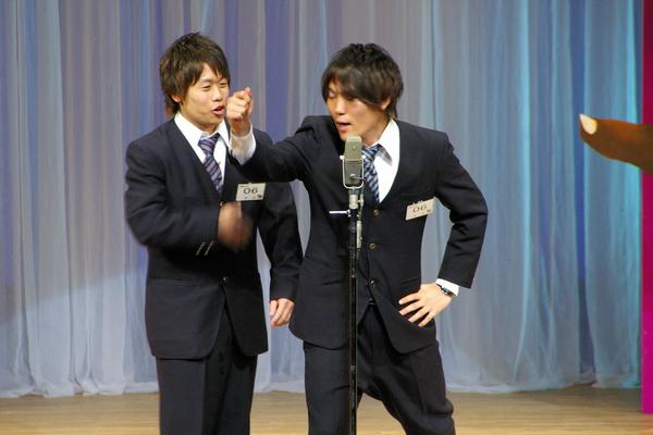 スーツを着た男性2人（音姫）がスタンドマイクの前で漫才をしている様子の写真