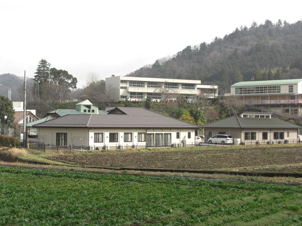 丘の上に小学校の校舎と体育館を背景に開設されたこげ茶色の屋根と緑色の屋根の2棟の幸の郷の写真