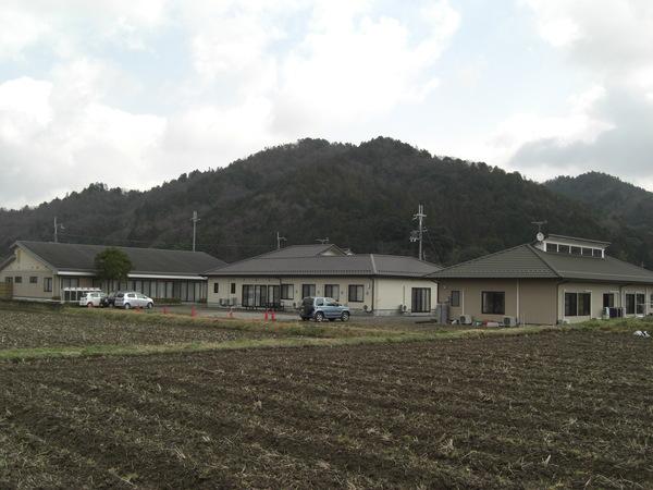 山並みを背景に開設されたこげ茶色の屋根2棟と緑色の屋根が並んで建つ福の郷の写真