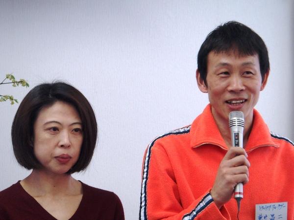 右側にオレンジ色の洋服を着ており、マイクを持って話をしている兼井 昌二さん,左側に紫の洋服を着ている奥様が写っている写真