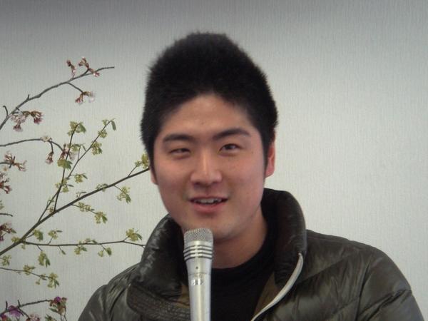 カーキ色のジャンパーを着ており、マイクを持って話をしている安達 鷹也さんの写真