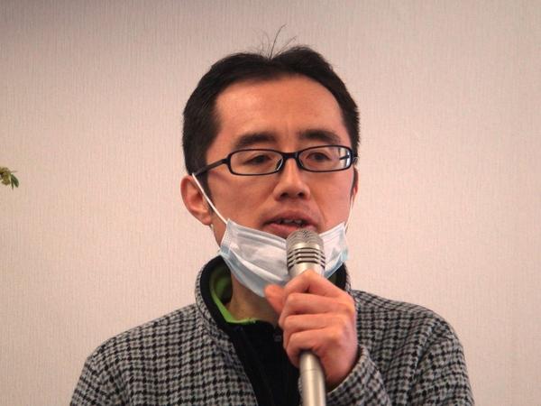 黒縁のメガネを掛けており、マスクをあごにずらして、マイクを持って話をしている初島 広治さんの写真