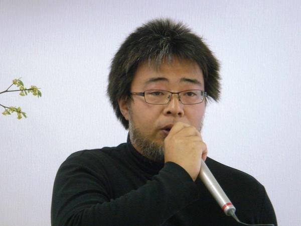 メガネを掛けており、黒い洋服を着ており、マイクを持って話をしている柿木 俊輔さんの写真