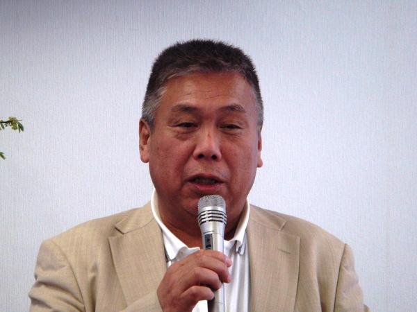 ベージュ色のジャケットを着ており、マイクを持って話をしている飯田 武志さんの写真