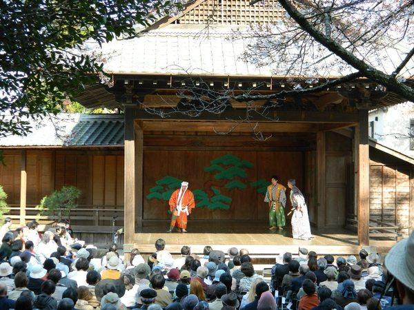 春日神社能舞台で白い布を被り、オレンジの衣装を着た方と黄色と緑の衣装を着た方とその横に白黒の衣装を着た男性が立っており、観客席から沢山の人々が見ている写真