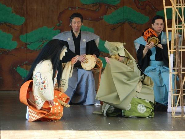 黒髪に白とオレンジの衣装を着た方と緑の衣装を着た方が手を顔に近づけており、その動きに合わせて後ろに座っている着物を着た2名の男性が小鼓を叩いている様子の写真