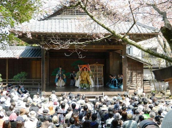 春日神社能舞台で黄色と緑の衣装を着た方が籠のようなものを持っており、右端には着物を着た方々が座っている、その狂言を観客席から見ている人々の写真