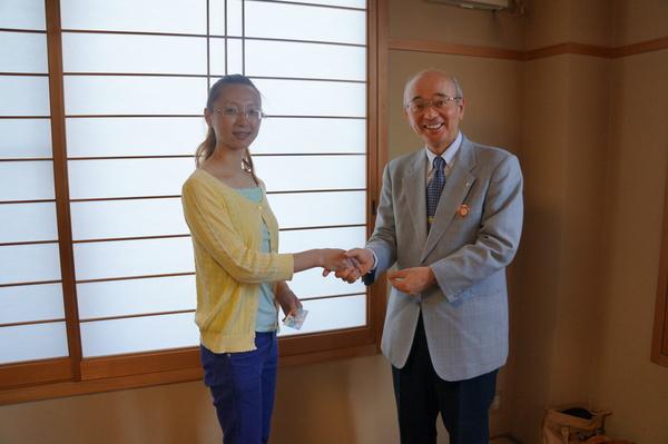 黄色のカーディガンにブルーのズボンの眼鏡の女性と市長が握手している写真