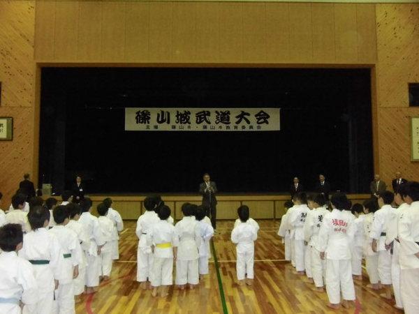 体育館の舞台の上に「篠山城武道大会」の横断幕が掛かっており、柔道に参加する選手達が整列しており、マイクの前で話をしてる男性が写っている写真