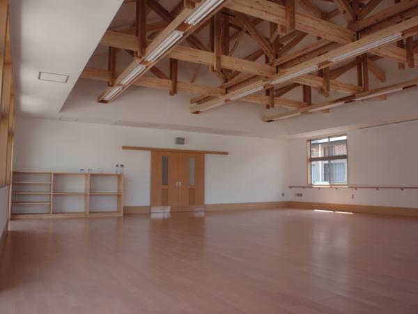 広い木の床が広がっていて、奥には棚とドアのあるリハビリ棟の室内が撮影されている写真