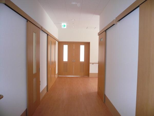 まっすぐと伸びた木の床の奥と横にドアがあるリハビリ棟の廊下の写真