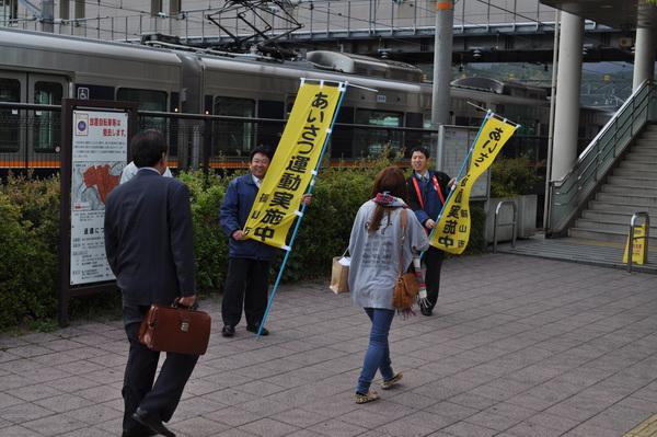 駅前で、市の職員男性2名が黄色の「あいさつ運動実施中」と書かれたのぼり旗を手に持って、通勤途中の女性と男性の市民に挨拶をしている様子の写真