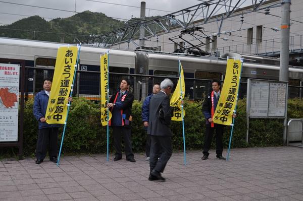 駅前で市の職員の男性4名が「あいさつ運動実施中」と書かれた黄色ののぼり旗を持って、通勤途中の白髪の男性に挨拶をしている様子の写真