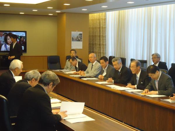 会議室にて出席者は向い合って座り、机に並べられた資料に目を通しながら、会議を進めている様子の写真