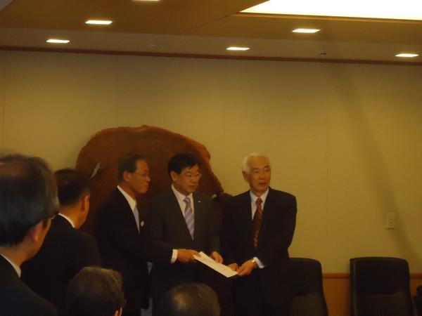 右にNHK松本会長がおり、紫色のネクタイを付けた男性と白髪の男性3人が白い紙を持っている様子の写真