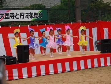 ステージ上で黄色、ピンク、紫、水色の衣装を着て頭には衣装と同じ色の大きなリボンを頭につけて両手を広げている女の子達の写真