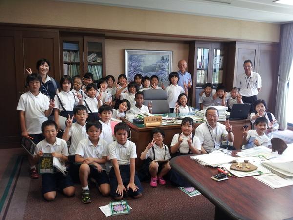 市長室の机の周りに集まりピースサインをする小学生、関係者、市長の写真
