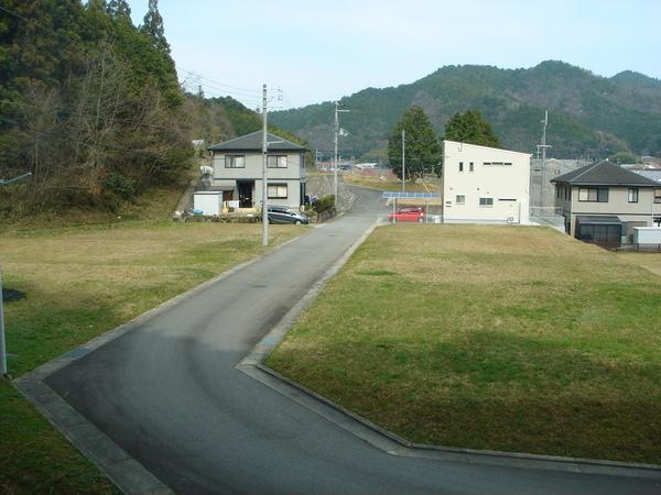 芝生が区画整理されている奥に白い2階建ての家と駐車された赤い車、道路反対側にグレーの2階建ての家に駐車されたグレーの車、その奥には山がみえる写真