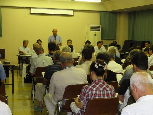 八上会場で前列の市長がマイクをもち立ちながら話をしているのを椅子に座り聞いている参加者の後ろ姿の写真