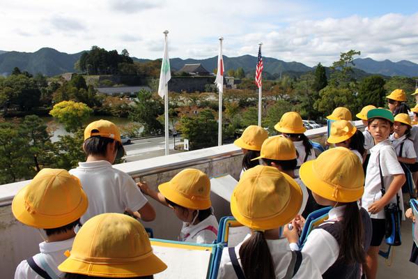 17名の黄色の帽子をかぶった小学生が市役所のベランダから外の様子を眺めている写真