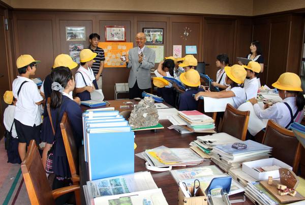 机の上にたくさんの資料が置かれていて、その周りに10名ほどの小学生が立っていて、市長が説明をしている写真