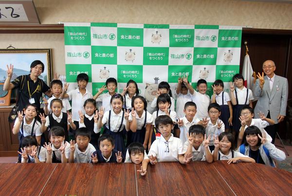 13名の男子児童と、13名の女子児童と手前に女性の先生、奥には男性の先生がいて、全員指で3を作って市長と記念撮影をしている写真