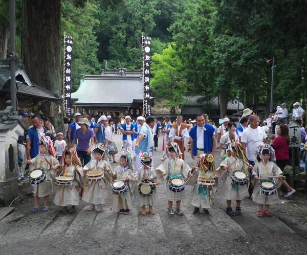 神社にて、白い衣装と被り物をした小さな子供達が横1列に並び太鼓を叩いており、その様子を見ている方々の写真